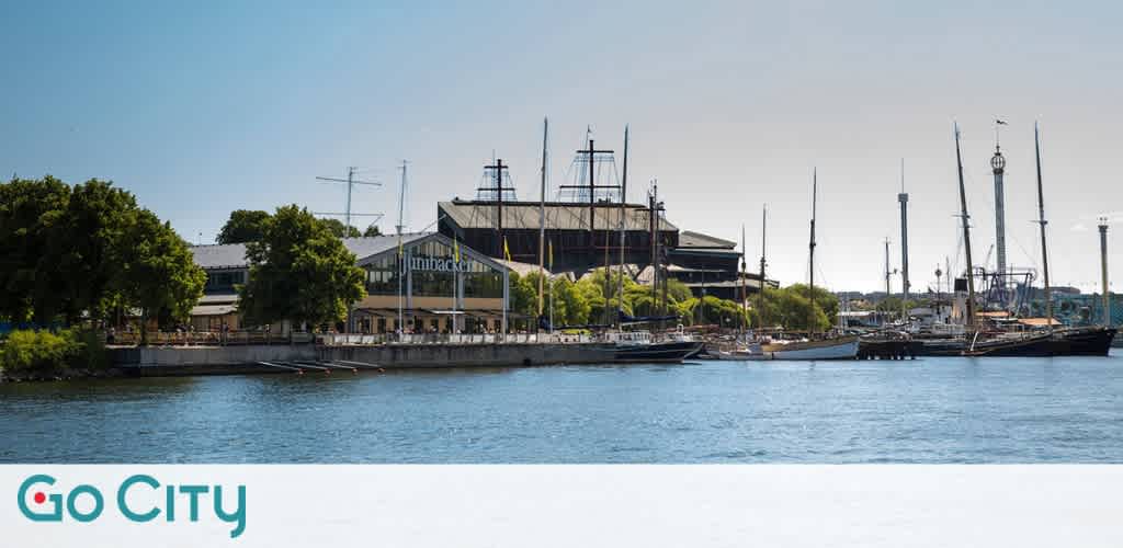 View of a waterfront with sailboats docked near a large building and green trees under a clear sky
