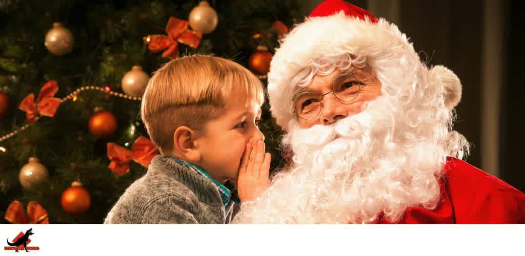 A young boy whispering to Santa Claus in front of a decorated Christmas tree.