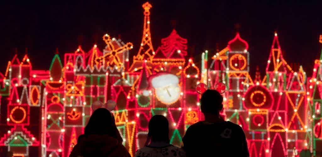 Three people watch a colorful, illuminated holiday display depicting a festive cityscape at night.