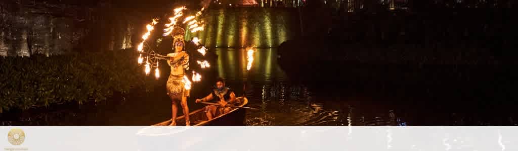 Two performers in traditional costumes perform fire dancing on a boat at night, with a lit bridge reflected in the water behind them.