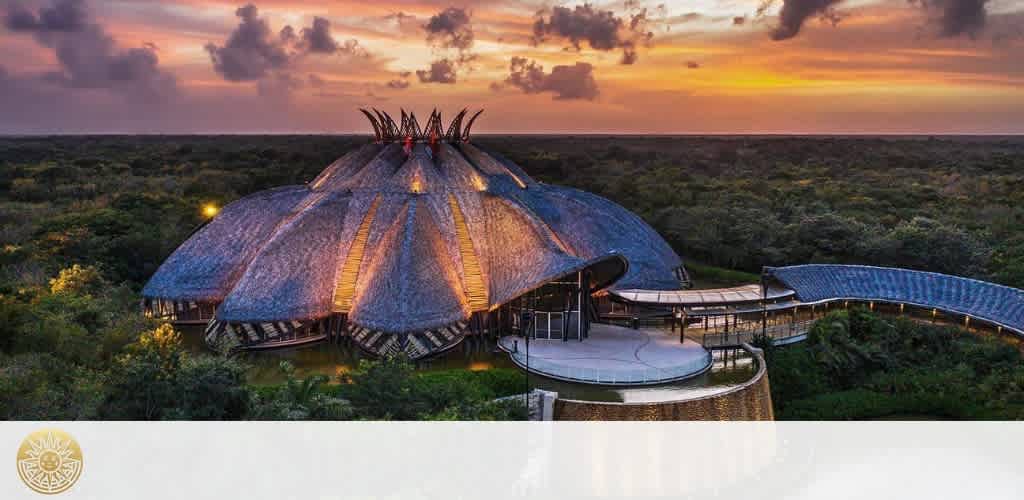 A large dome-shaped building with a thatched roof and surrounding walkways set in a lush green landscape during sunset.