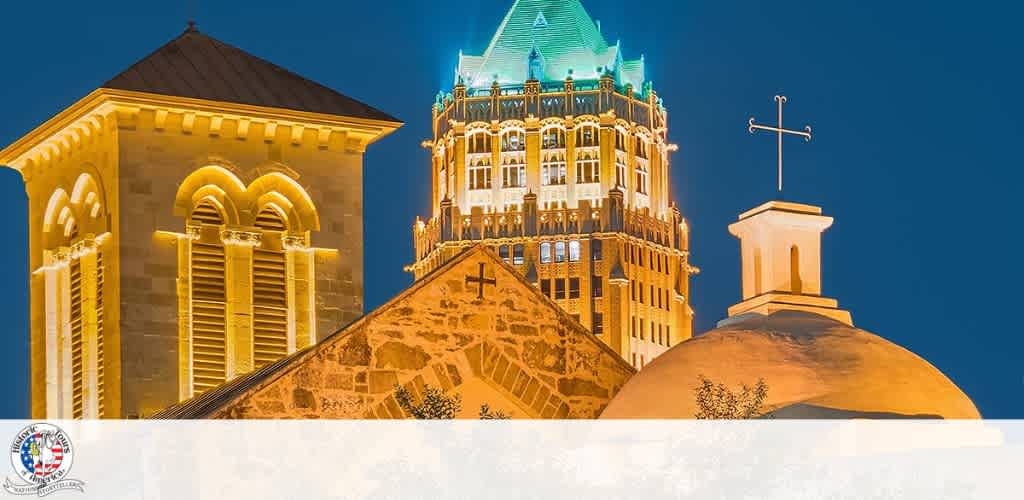 Night view of illuminated historic church towers with cross and clock tower against a dark blue sky