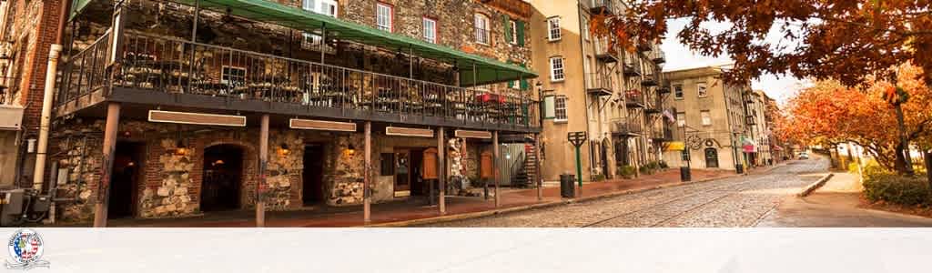 A cobblestone street lined with multi-story brick and stone buildings with balconies and fall trees with orange and yellow leaves