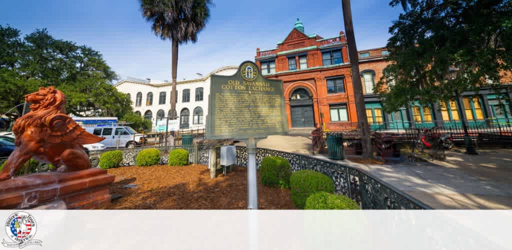 Historic building with a red brick facade, a historical marker, green trees, and a lion statue in front, surrounded by a black decorative fence and a sidewalk.