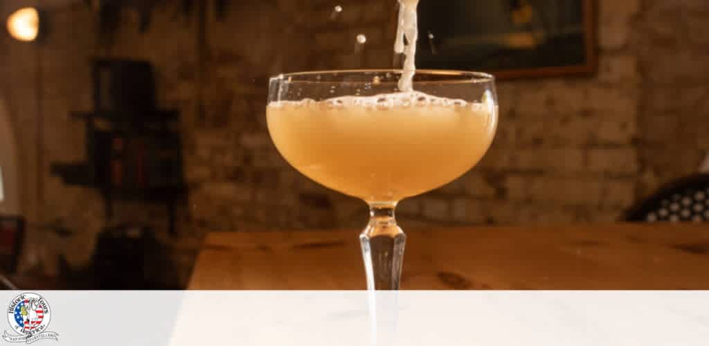 A cocktail glass with a stem filled with a light-colored drink, being poured inside a cozy brick-walled room.