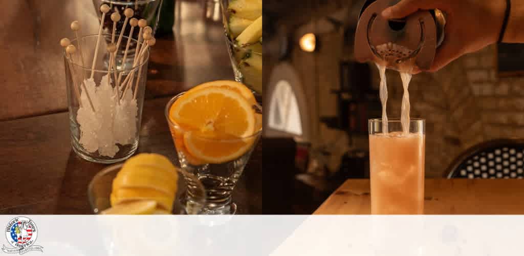 A hand pours a beverage into a glass, with fresh orange slices and cocktail accessories on a wooden table in a cozy bar setting.