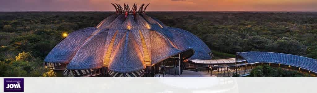 A large, thatched-roof building with multiple peaks and surrounding greenery, illuminated at sunset with warm, colorful skies overhead.