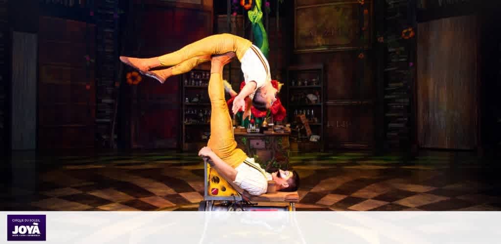 Two performers in colorful costumes are balancing during a circus act on a decorated stage with wooden walls and flooring.