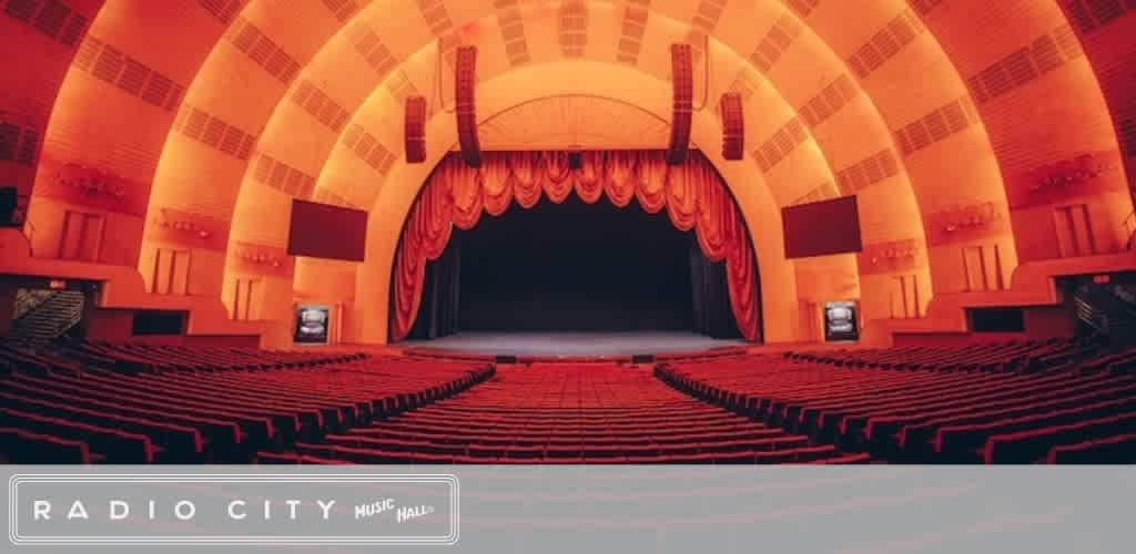 An empty theater with red seats, a stage with red curtains, and large screens on either side in a grand, curved auditorium with warm lighting.