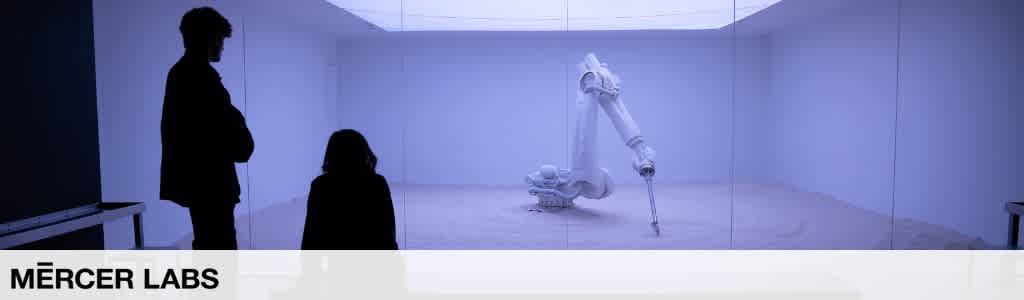 A person stands and a child sits in front of a robotic arm exhibit in a white, high-tech lab environment at Mercer Labs.