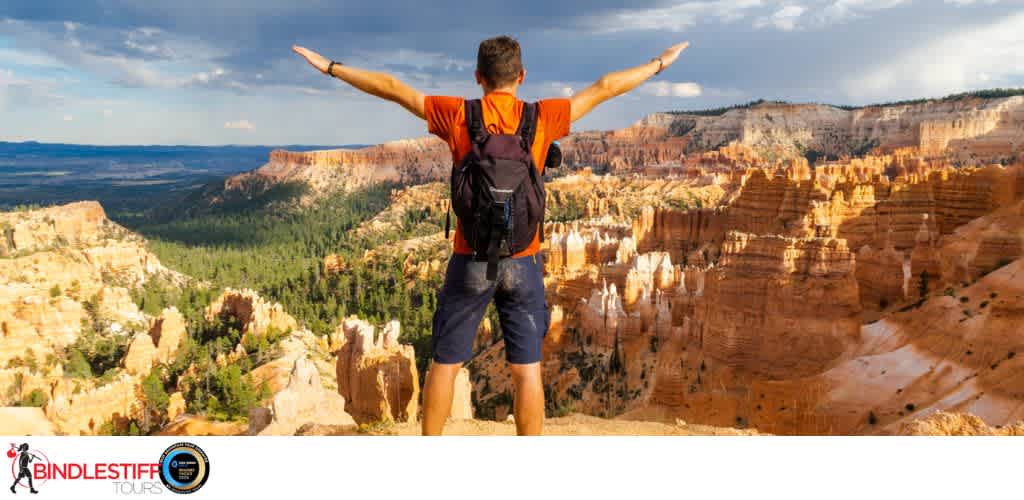 A person stands with arms outstretched overlooking a canyon landscape with cliffs and green trees under a partly cloudy sky