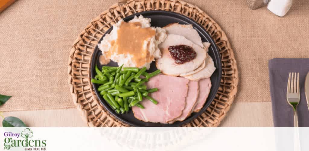A meal of turkey, ham, green beans, and mashed potatoes with gravy on a black plate.