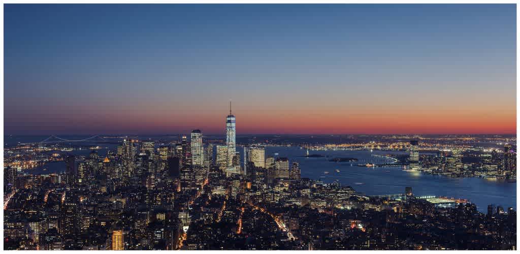 A city skyline at sunset with illuminated buildings and a river reflecting the sky's fading light in the background
