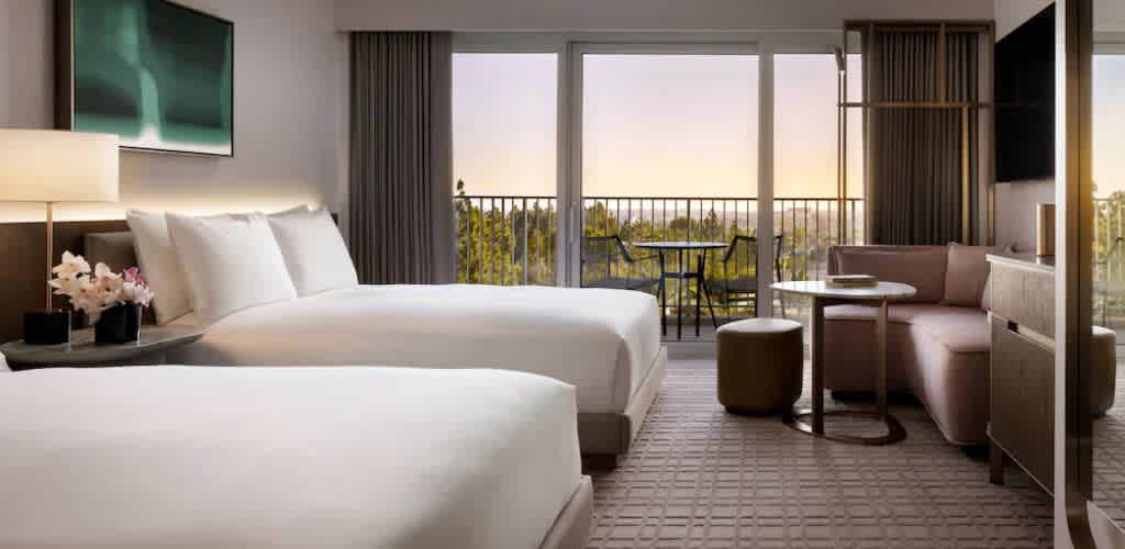 Contemporary hotel room with two beds, a seating area, a balcony with outdoor furniture, large windows, and scenic views at sunset