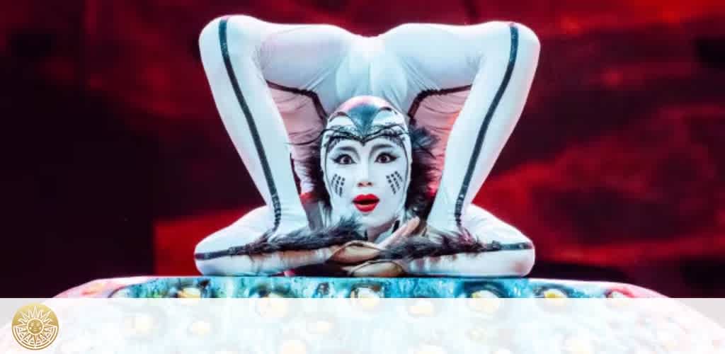 A person with white face paint and black face markings, wearing a white costume with black accents, is performing with an expressive facial expression.