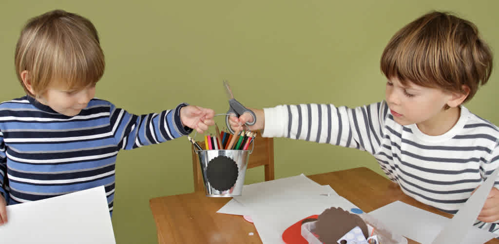 Two children with striped shirts are exchanging objects at a desk with art supplies against a green background
