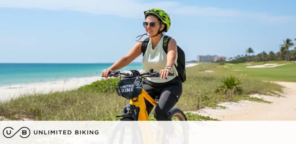A woman rides a yellow bicycle along a coastal path with ocean and green landscape in the background. She wears a helmet and sunglasses.