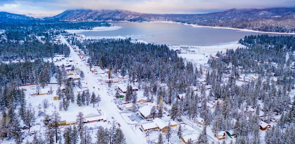 Aerial view of a snow-covered lakeside town with houses surrounded by trees and a frozen lake in the background during winter.