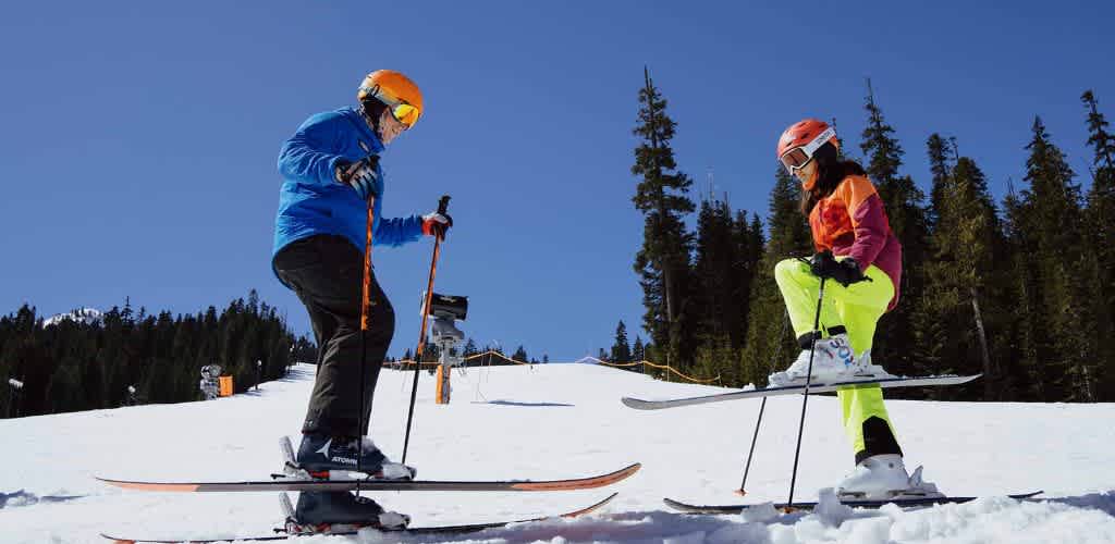 Two children wearing colorful ski gear and helmets ski on a snow-covered slope with a clear blue sky and pine trees in the background