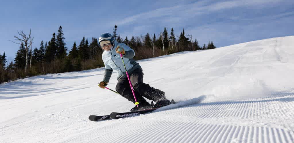 A person skiing downhill on a snowy slope with a backdrop of trees and a clear blue sky
