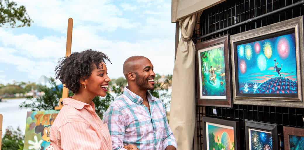 Two people happily viewing colorful framed artwork displayed outdoors at an art fair or exhibition