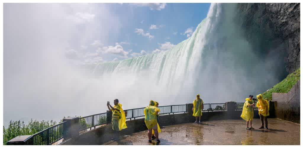 Visitors wearing yellow rain ponchos stand on a viewing platform near Niagara Falls with mist and water spray in the background