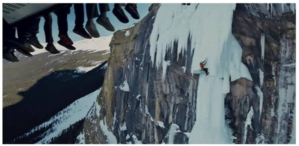 A person ice climbing on a frozen waterfall with a mountain backdrop, seen from below a ski lift overhead.