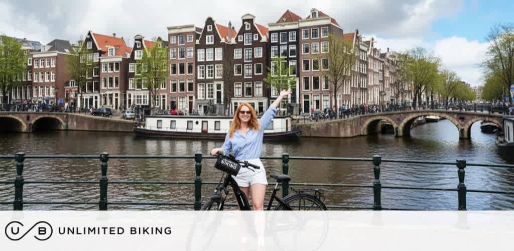 A woman with red hair and sunglasses standing next to a bicycle by a canal with historic buildings and a bridge in the background