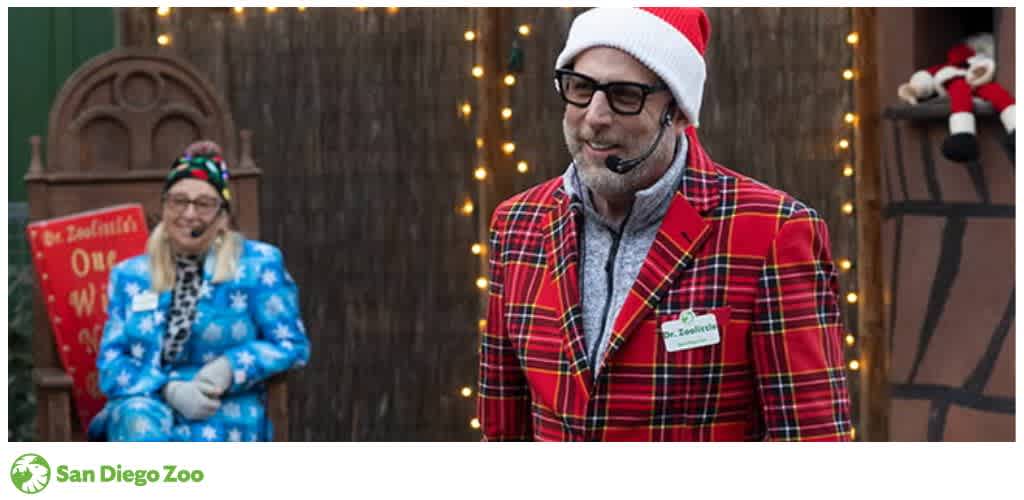 Two people in festive attire with microphones, one standing in a plaid jacket and the other seated in a blue snowflake suit, at a decorated event.