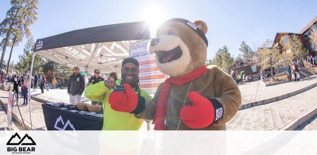 Costumed bear mascot and person posing at Big Bear Mountain Resort with a tent and lodge in the background.