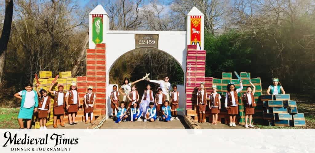 Children in uniforms standing in front of a cookie box archway outdoors, surrounded by trees.
