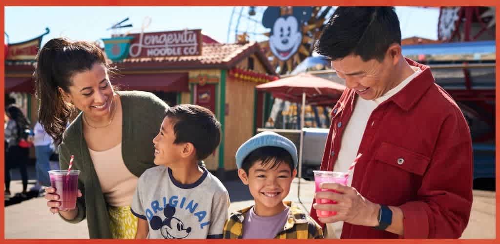 A smiling family enjoying drinks at an amusement park with rides and colorful signage in the background.