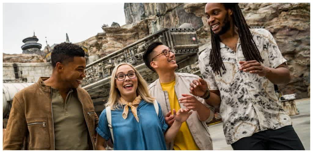 Group of diverse friends smiling and talking outdoors with a rocky background and spaceship replica.