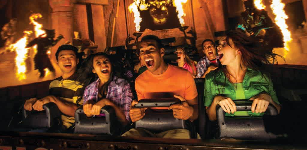 Six people are riding a roller coaster with flames in the background, expressing excitement and thrill during the ride.