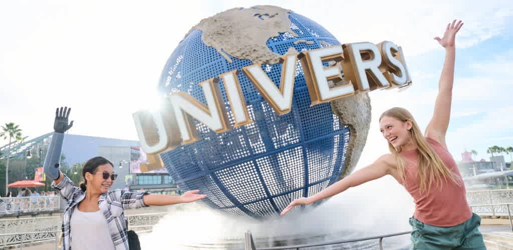 Two smiling women interact with a large globe displaying the word UNIVERSE at a theme park. The scene is lively with sunny weather and palm trees in the background.