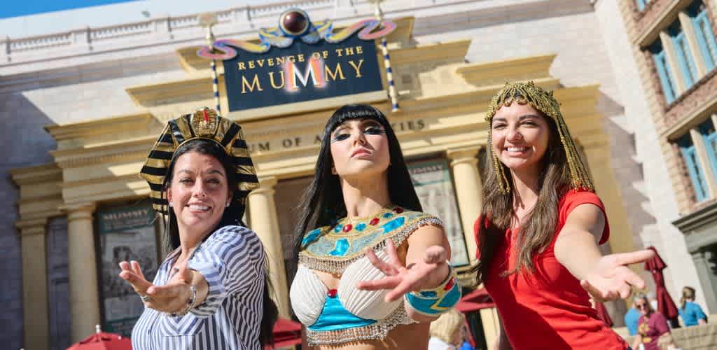 Three women in costumes pose playfully in front of the Revenge of the Mummy attraction at a theme park