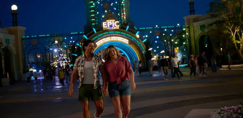 Two women walk happily at night outside Epic Universe amusement park entrance surrounded by colorful lights and other visitors.