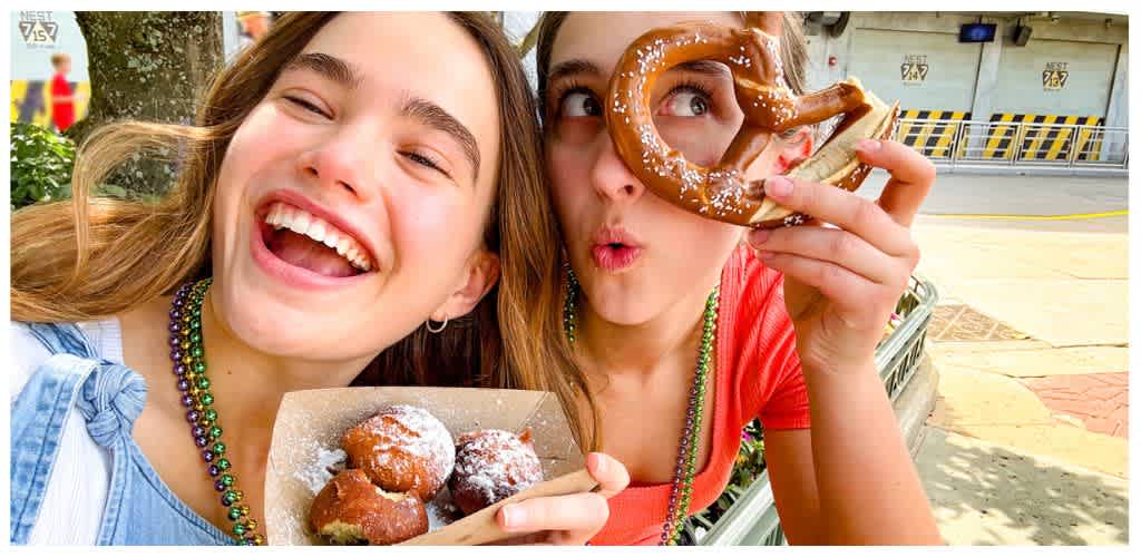 Two smiling women enjoying baked goods outdoors, one holding a plate of donuts and the other holding a pretzel.