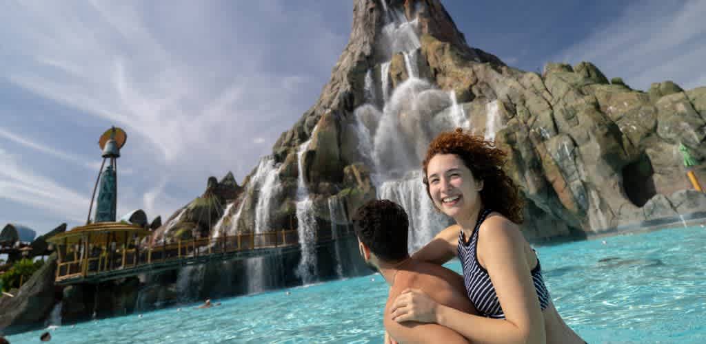 Two people are happily embracing in a swimming pool with a waterfall and rocky mountain in the background on a sunny day.