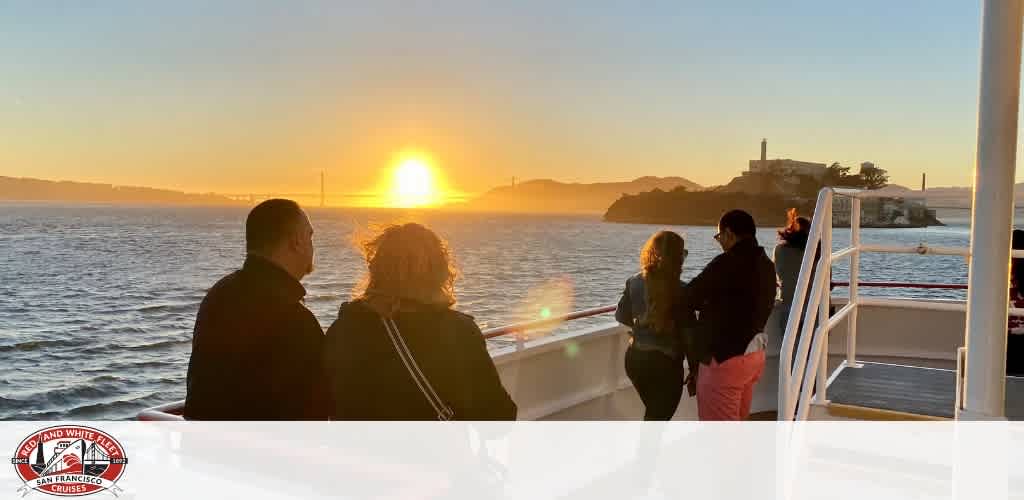 People on a boat deck watching a sunset over a body of water with a bridge and an island in the distance.