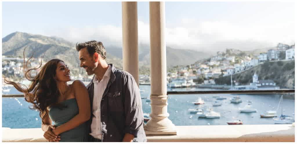 A happy couple standing on a balcony overlooking a harbor with boats and hills in the background
