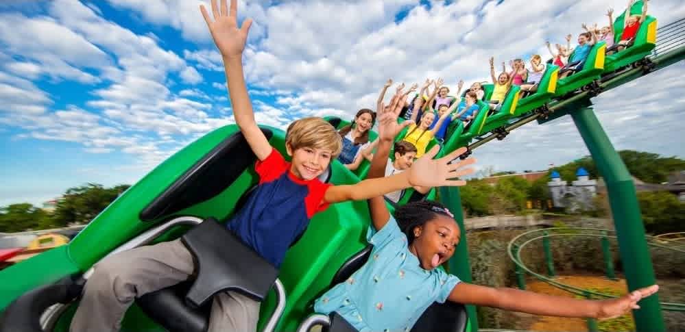 Children are enjoying a roller coaster ride outdoors with smiles and arms raised, experiencing fun and excitement on a bright, sunny day.