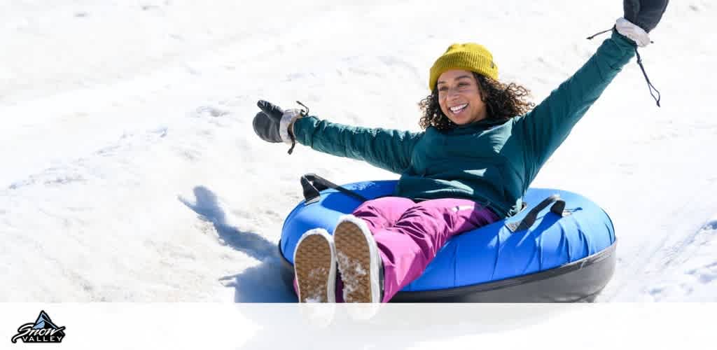 Woman smiling and sitting in a blue snow tube on snow with arms raised, enjoying a winter activity outdoors