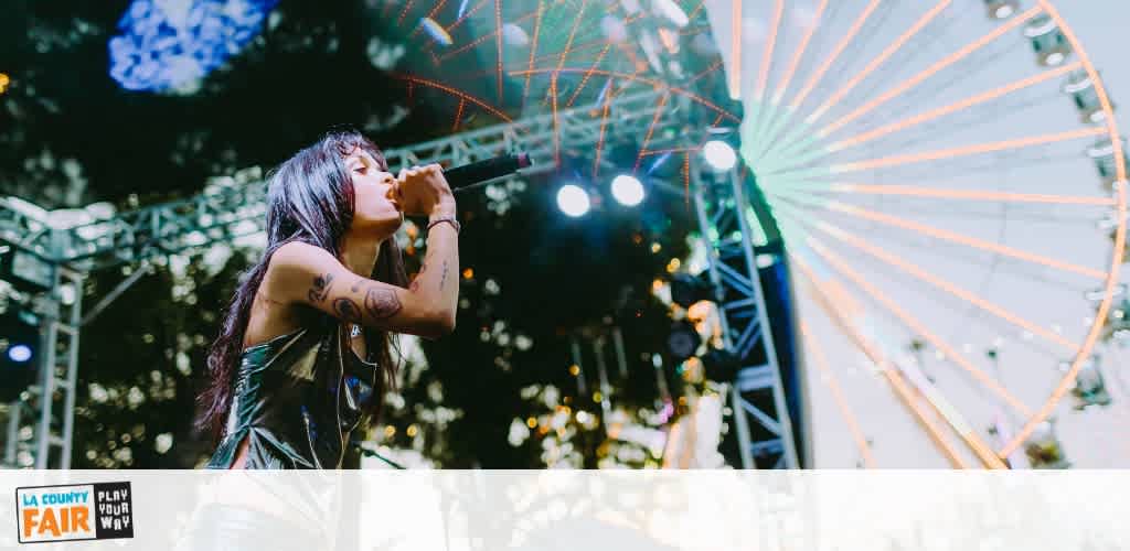 A woman singing on stage at a fair with a Ferris wheel in the background during an outdoor evening event.