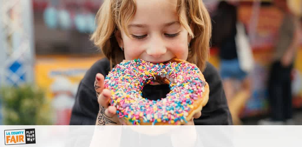 A young girl with blonde hair enjoys a colorful sprinkled donut at an outdoor fair