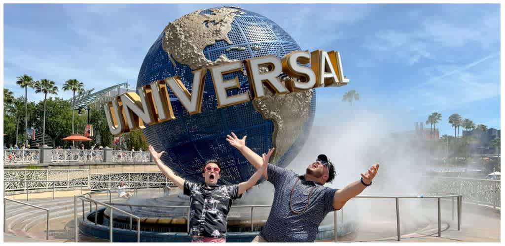 Two people pose excitedly in front of the Universal Studios globe at the entrance of the theme park with blue sky and palm trees in the background.