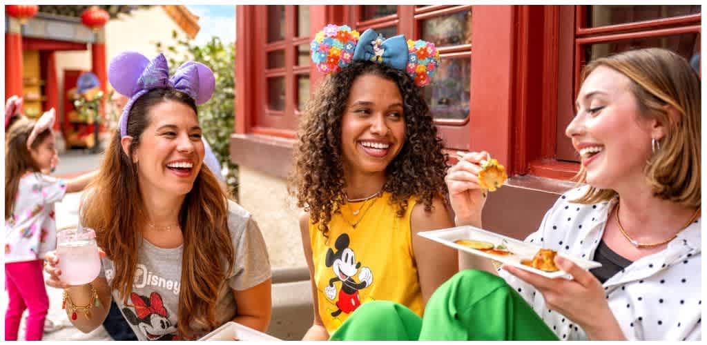 Three women laughing and enjoying snacks outside at a celebration wearing Disney themed headbands and casual clothing.