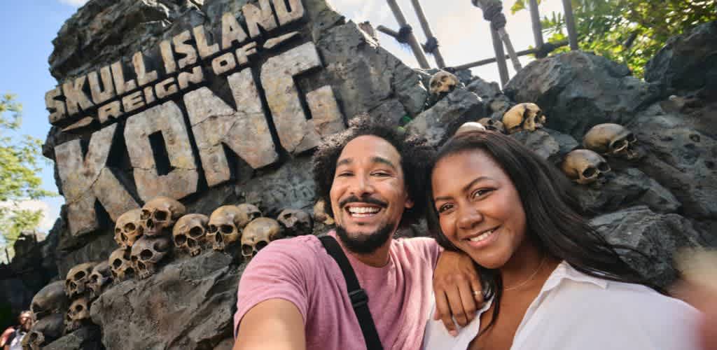 A smiling couple taking a selfie in front of Skull Island Reign of Kong sign with skulls and rocks on display at an amusement park