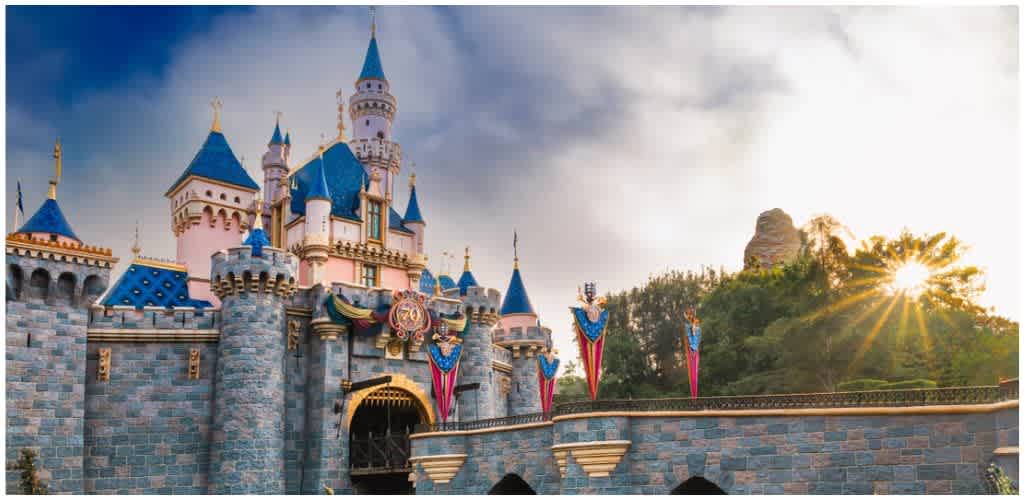 Colorful Disney castle with pink, blue, and gold accents, surrounded by a stone wall and lush greenery under a bright, partly cloudy sky.