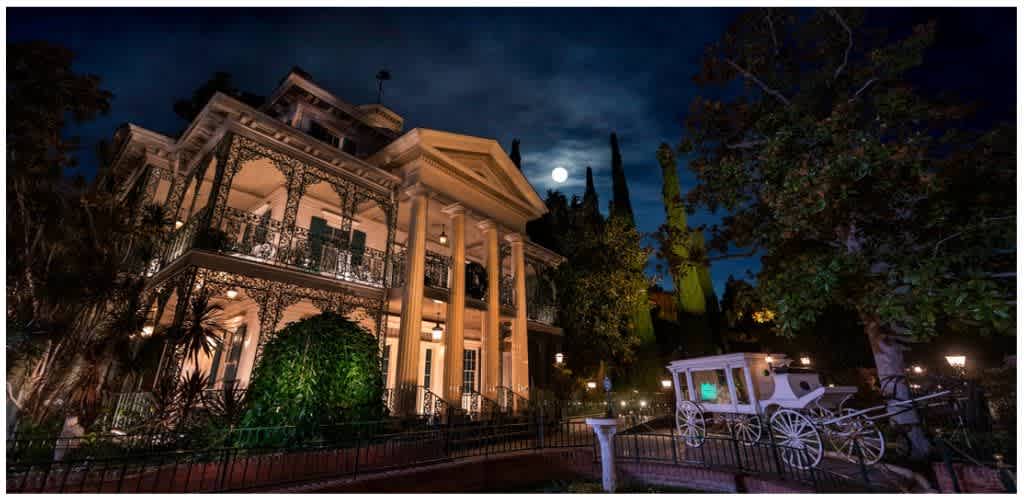 Historic mansion illuminated at night with full moon in the sky and a white carriage in front surrounded by trees and garden lights